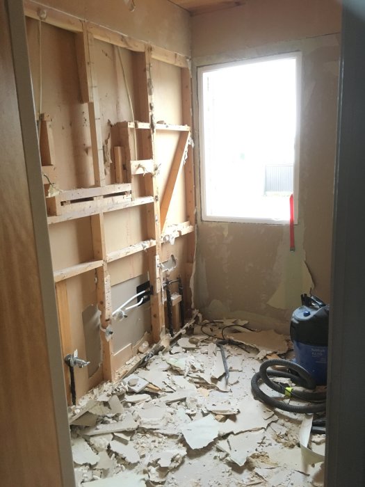 Renoveringsrum med borttagen gipsvägg som visar trästommar och rör, med spår av byggavfall på golvet.