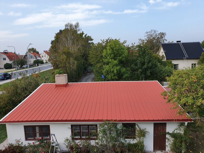 Rött tak på en villa med skorsten, synlig från ovan i dagsljus, omgiven av grönska och gata.