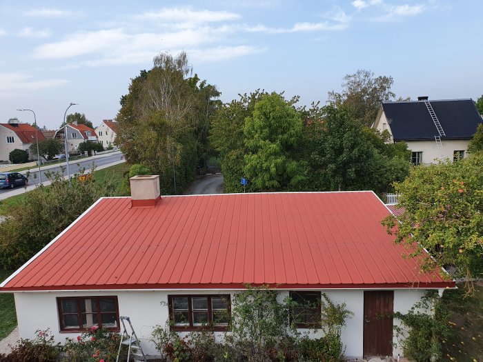 Röd takpapp på en villa, omgivning med andra hus och grönska, himmel och skorsten synlig.