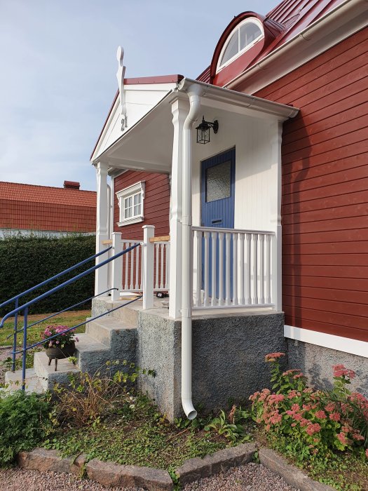 Entré till rött trähus med en blå ytterdörr, vit veranda, och en rund fönsteröverdel.