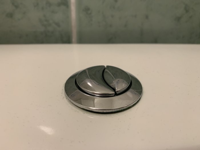 Rund toalettspolknapp med två knappar mot en grönaktig vägg.