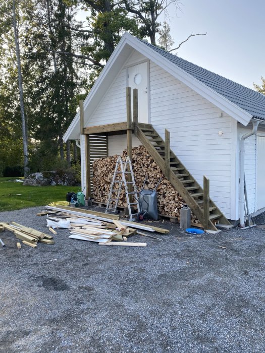 Vit uthusfasad med vedstapel, stege, och lösa träplankor på marken under konstruktion.