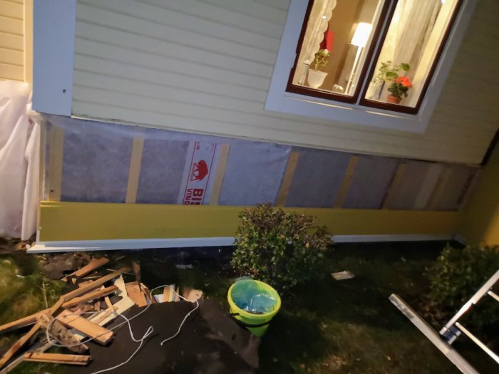 Husvägg under renovering med delvis målade panelbrädor och oisolerade sektioner synliga vid skymning.