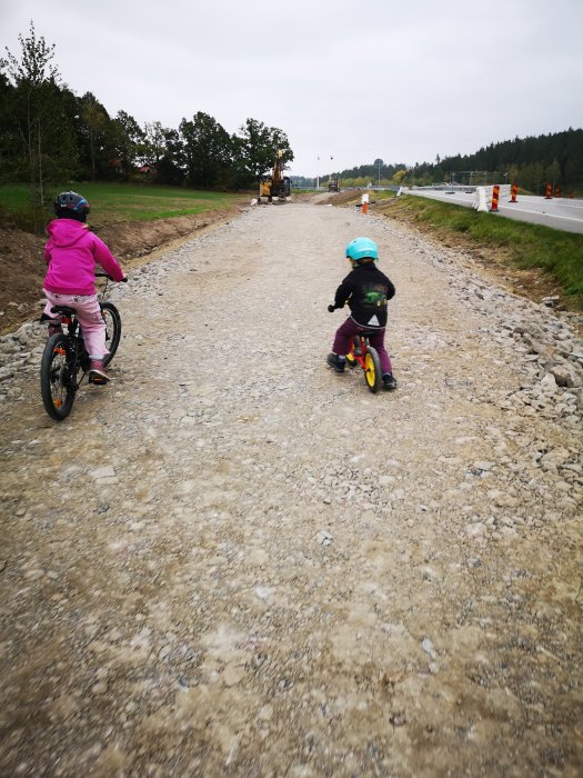 Två barn på cyklar på en grusig, nybyggd cykelväg med anläggningsmaskiner i bakgrunden.