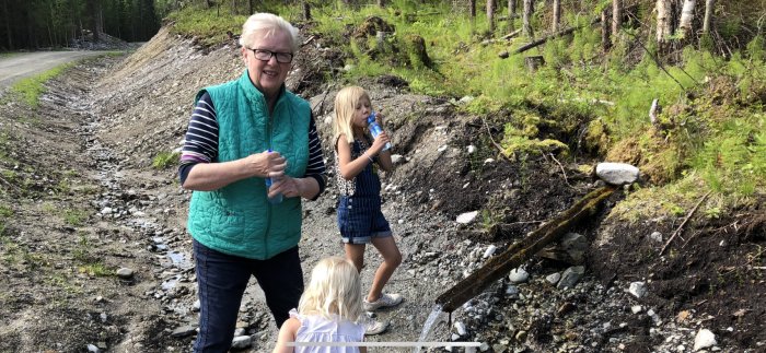 Två barn och en äldre person med vattenflaska utomhus på en grusig stig vid skog.