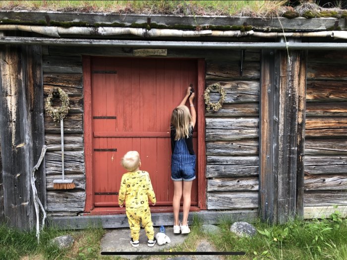 Två barn framför gammal seterstuga, pojke i pyjamas & flicka öppnar dörren.