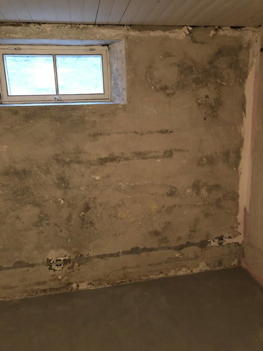 En ojämn betongvägg med fläckar av borttagen färg och små hål, under ett fönster i ett renoveringsprojekt.