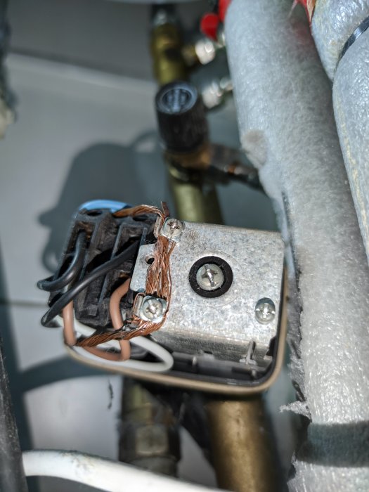 Elektrisk kopplingslåda med flera hopvridna kablar och en skruvansluten anordning, mot bakgrund av rör.