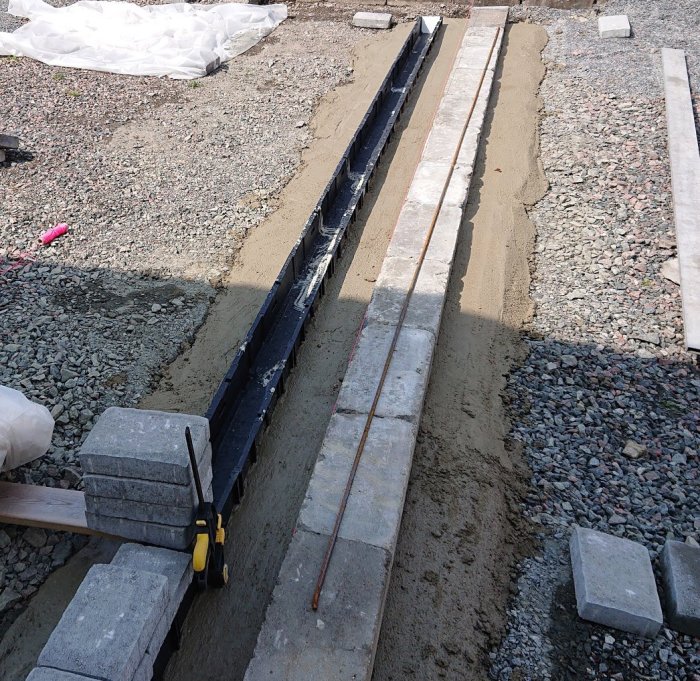 Installation av dagvattenavrinning med en lång smal grävd ränna och betongfundament vid en plattyta under arbete.
