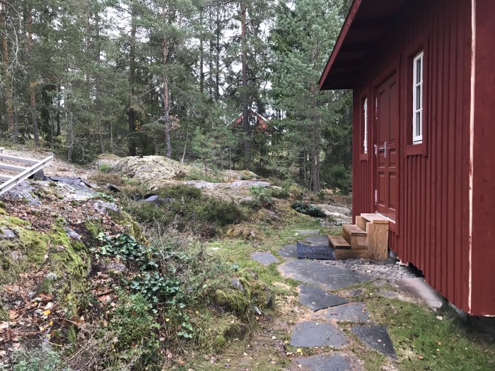 Röd trähusfasad med dubbla ytterdörrar och en tillfällig trätrappa i ett skogsläge nära ett berg.