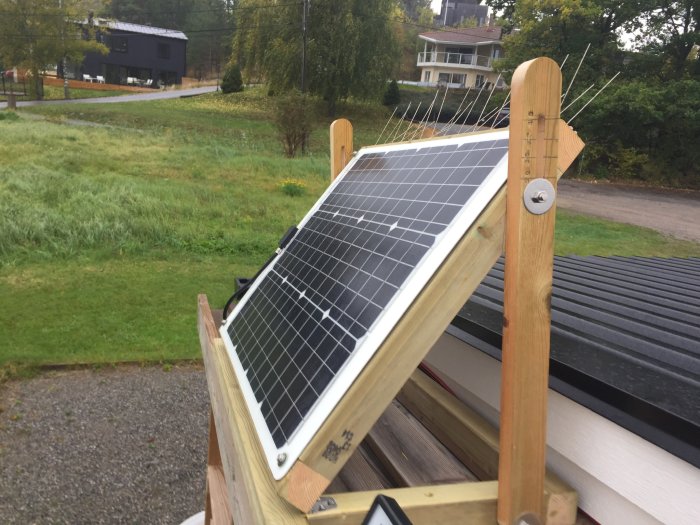 Solpanel installerad på träställning med gräsbevuxna fält och hus i bakgrunden.