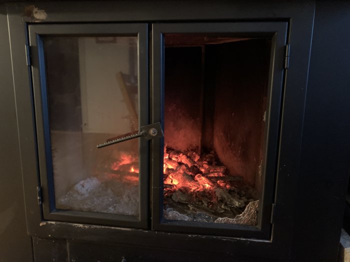 Öppen plåtkamin med glödande ved och delvis suddigt glas, där en ny tätningslist har monterats.