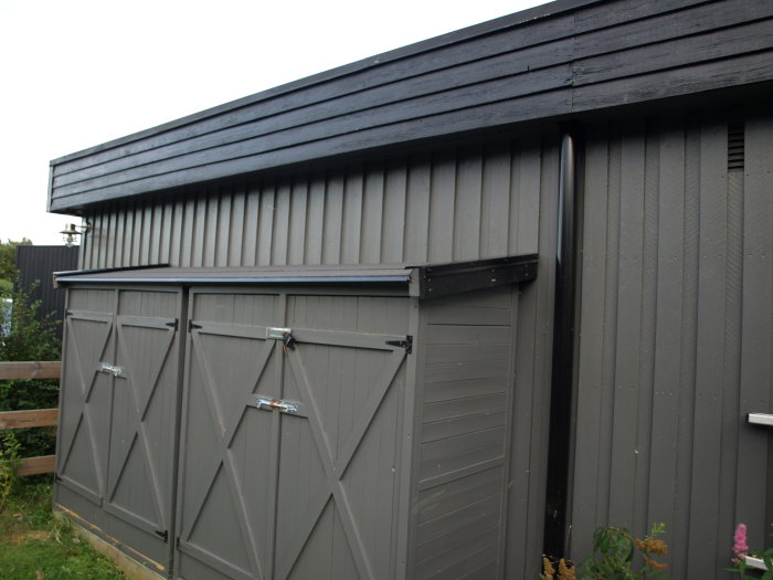 Mörkgrå låsbar redskapsbod för förvaring av färgburkar och sopsäckar, med dubbla dörrar och vinkelrätt tak.