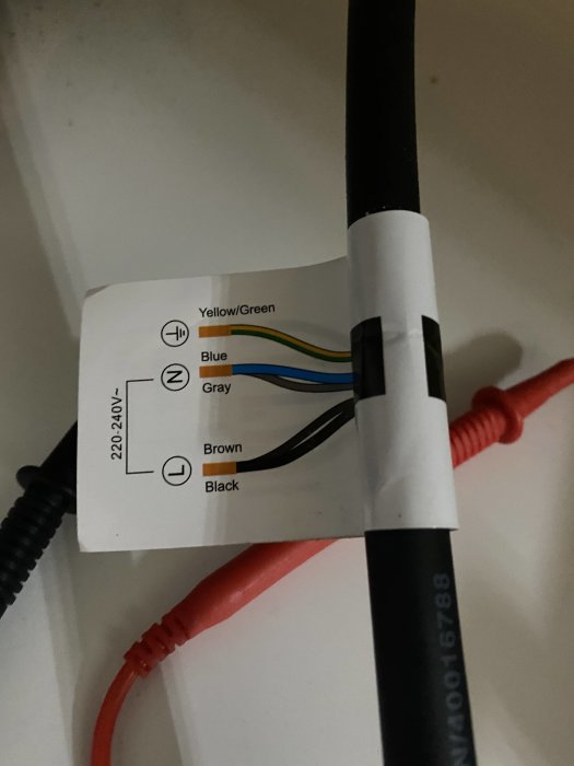 Etikett som visar kabelfärgskoder och anslutningar för en 220-240V installation mot en svart elektrisk kabel.