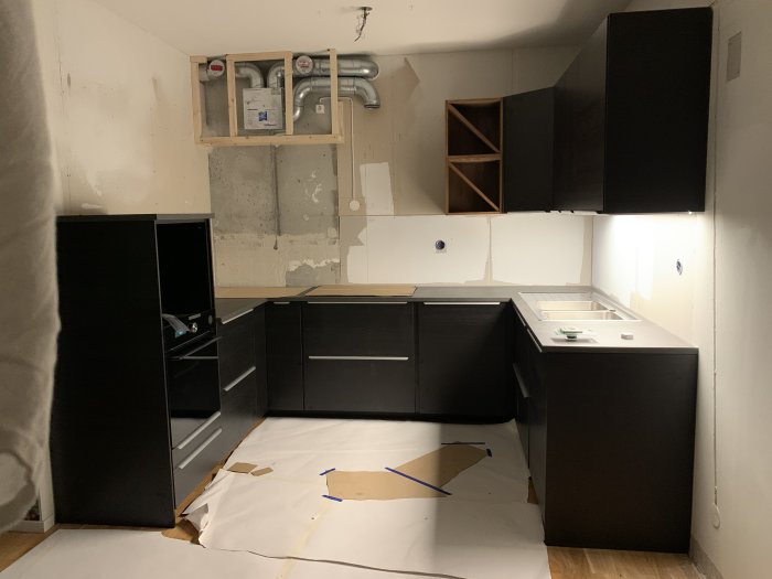 Ej färdigt kök med flyttat fläktdon i hörn och synliga rördragningar, samt skåp utan luckor och kakel.