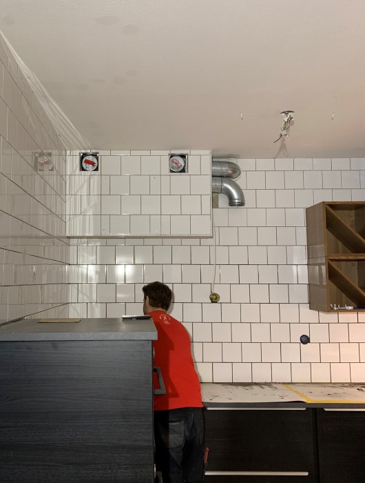 Kök under renovering med en person som justerar fläktdonet i en vit kaklad vägg med synliga rör och en öppen låda.