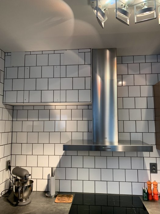 Kökshörn med ombyggda rördragningar och inbyggd fläkt ovanför induktionshäll, vit kakelvägg och belysning.