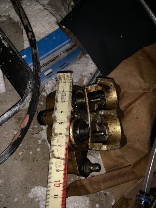 Bromsok med mätmått som visar ca 4,5 cm avstånd mellan skruvfästen i en rörig verkstadsmiljö.
