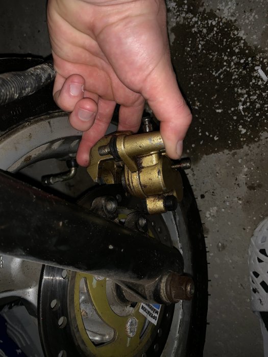 Hand som håller i ett gyllene bromsok monterat på ett motorcykelhjul, med fokus på fästpunkterna.