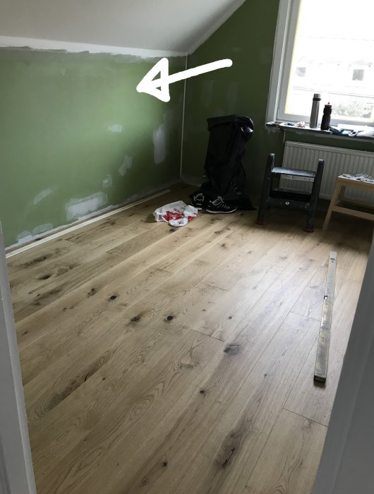 Rum under renovering med trägolv och väggar delvis spacklade, vit pil pekar på vägg.