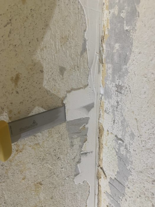 Vägghörn med gammalt spackel och rester av brun tapet, spackelkniv används för borttagning.