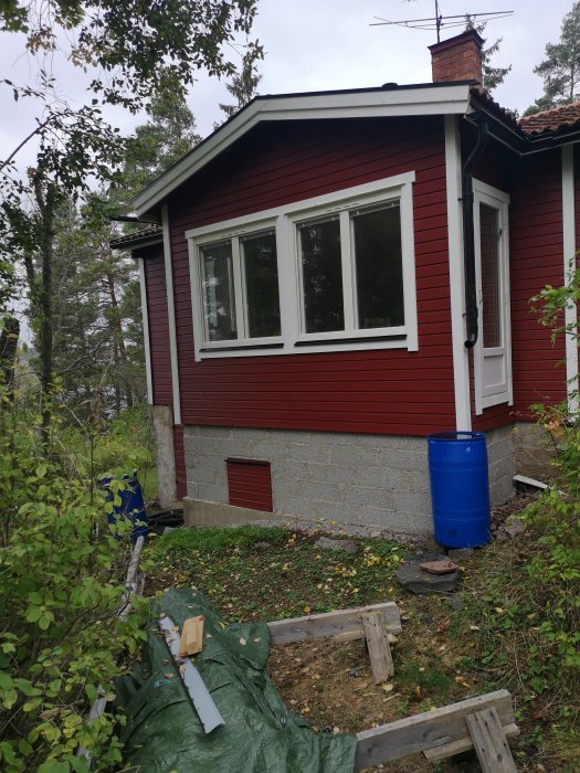 Rödmålat hus med vita fönster, vindskivor och små luckor i grunden, byggmaterial i förgrunden.