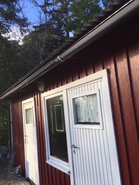 Ytterväggen på ett hus med röda plankor, vita knutar, två dörrar och ett fönster som ska ersättas med pardörrar och en regel.