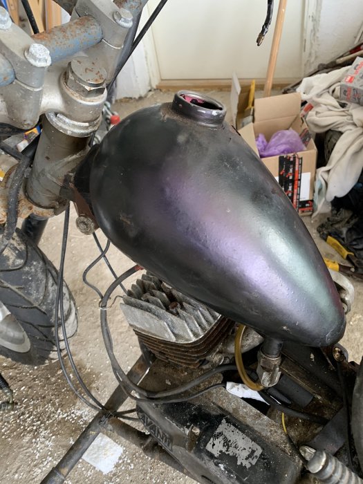 Närbild på en motorcykels bensintank och motor i en verkstad, tanken är otydligt märkt med volym.