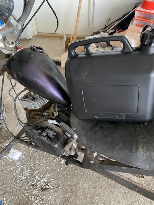 Motorcykeltank bredvid svart plastdunk i ett garage, verktyg och byggmaterial i bakgrunden.