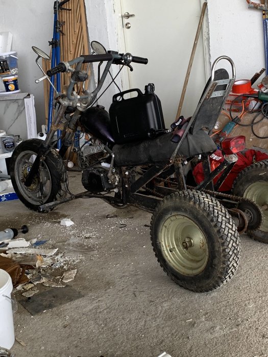 Egenbyggt trehjuligt fordon i en garageverkstad med diverse verktyg och föremål i bakgrunden.
