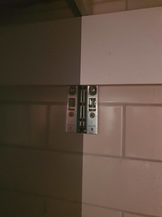 Metallbeslag mellan två vita ytor, ej fullt synlig bakom vit platta.