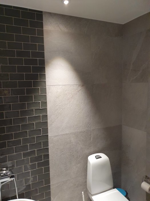 Liten badrumshörna med högt i tak, gråa kakelväggar och mörkt kakel vid spegeln ovanför toaletten.
