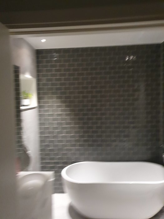Ett litet badrum med mörka kakelväggar, fristående badkar och toalett, suddig bild.