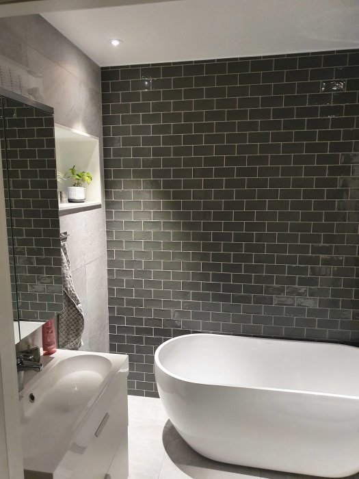 Modern fristående badkar och handfat i ett badrum med mörka kaklade väggar och inbyggd hylla.