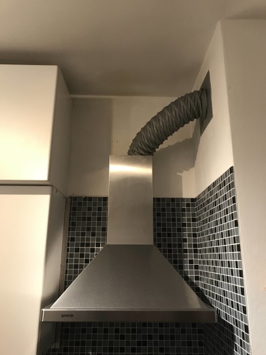 Köksfläkt ansluten till flexibel ventilationsslang i ett kök med svart kakel.