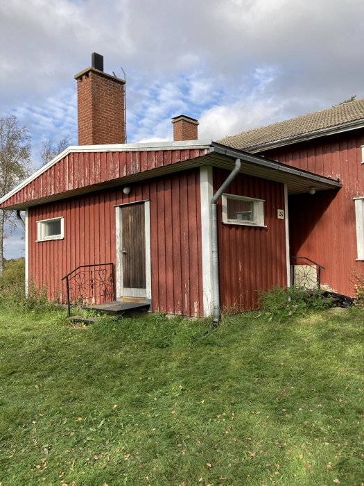 Rödmålad tillbyggnad med litet fönster och enkel dörr mot en gräsmatta, ger ett intryck av 1970-talet.