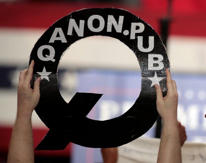 Person håller upp en stor Q-symbol med texten "ANON.PUB" mot en bakgrund med amerikanska flaggan.