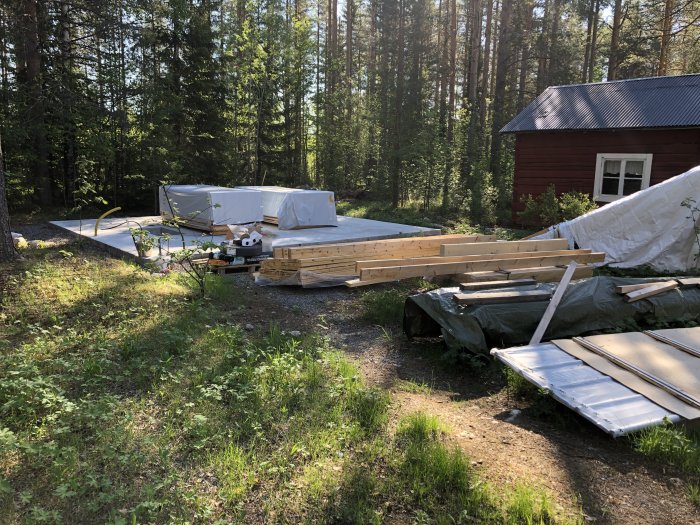 Betongplatta med staplade byggmaterial skyddade under presenningar i en skogsglänta, nära ett rött hus.