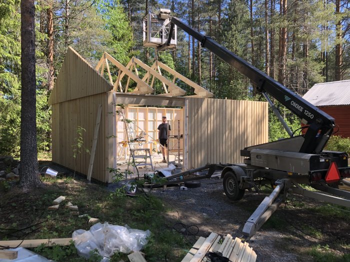 Byggprocess av snickarbod med takstolar som lyfts av en skylift, omgivet av skogsmiljö.