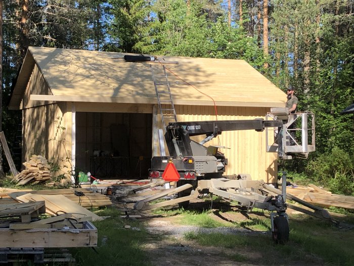 Halvfärdig snickarbod under konstruktion med takstolar, synliga väggblock och en person i en skylift.