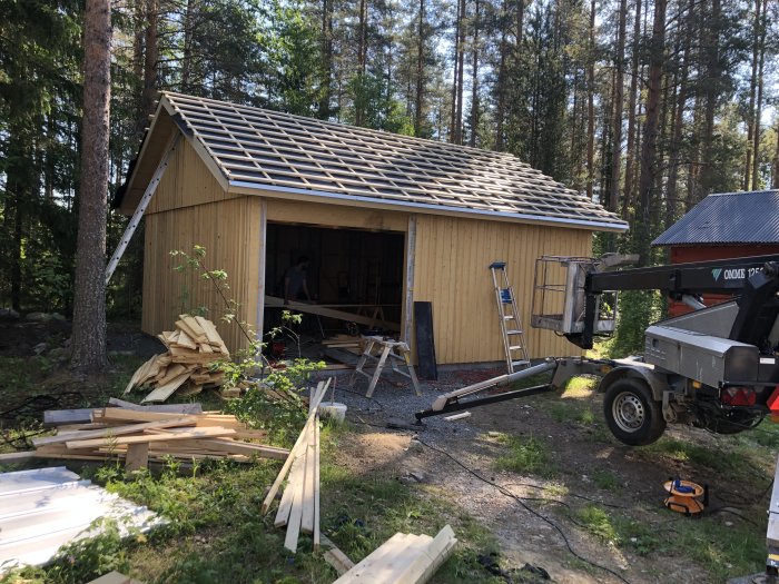 Förrådsbyggnad under uppförande med oskyddat tak, byggmaterial runtomkring och en skylift.