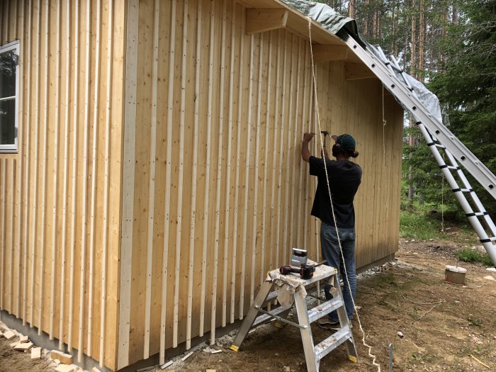 Person som målar en nybyggd träförrådsfasad, med stege och skog i bakgrunden.