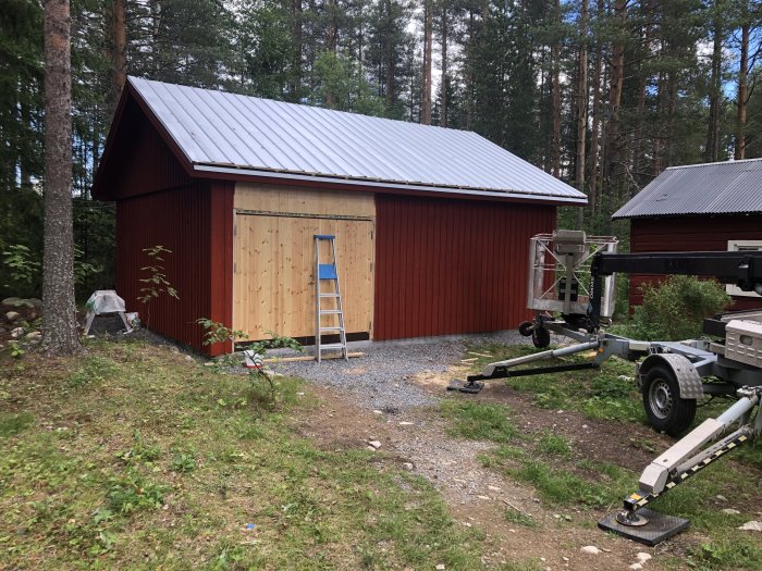 Nybyggd snickarbod med röd panel, faluröd färg och pannplåtstak, vid skogsbryn med stege upp till dörren.