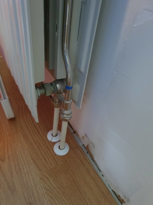 Radiatorventil vid golv med synligt rörsystem och ventilhus, intill en vit vägg och på ett parkettgolv.
