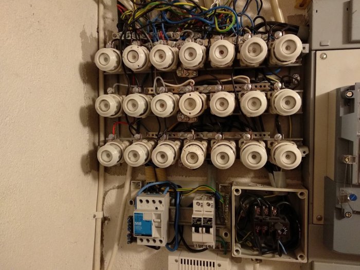 Elektrisk panel med många säkringar, röriga kablar och en effektvakt, i behov av omdaning.