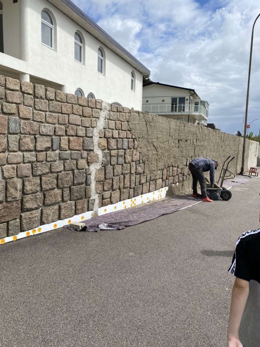 Nyputsad mur med ojämn naturstenstopp nära vit byggnad, person fäster plåtar, reflexskyltar på marken.