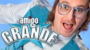 Person i turkos dräkt med texten "amigo GRANDE", glasögon och leende mot kameran.