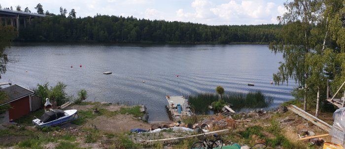 Utsikt från terass över sjö med brygga, båt och rött hus vid strandkant, med skog i bakgrunden.