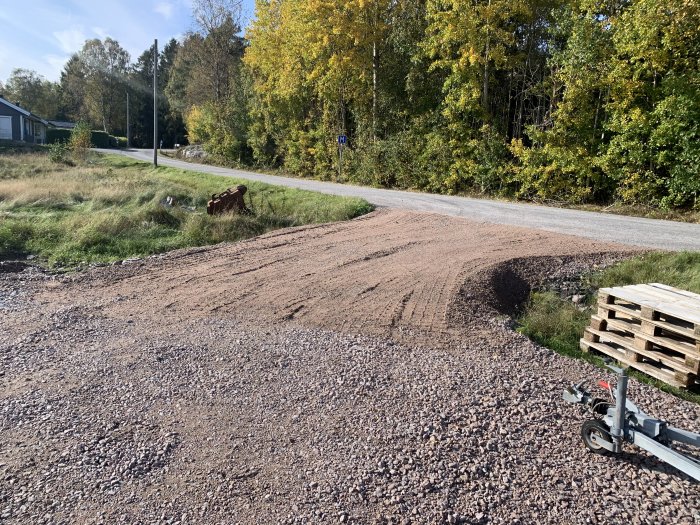 Grus spritt över infarter med materialhög intill garage och tomtpallar i förgrunden, landsväg och höstträd i bakgrunden.