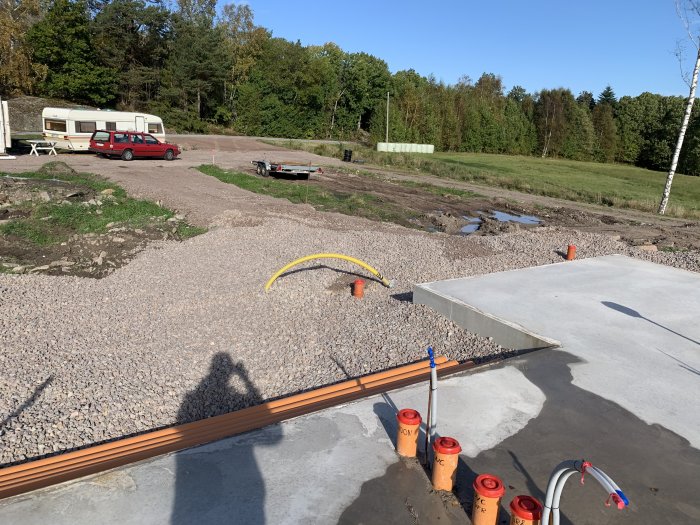 Tomt med utspridda grus och betongplatta nära anlagd infrastruktur och parkerade fordon.